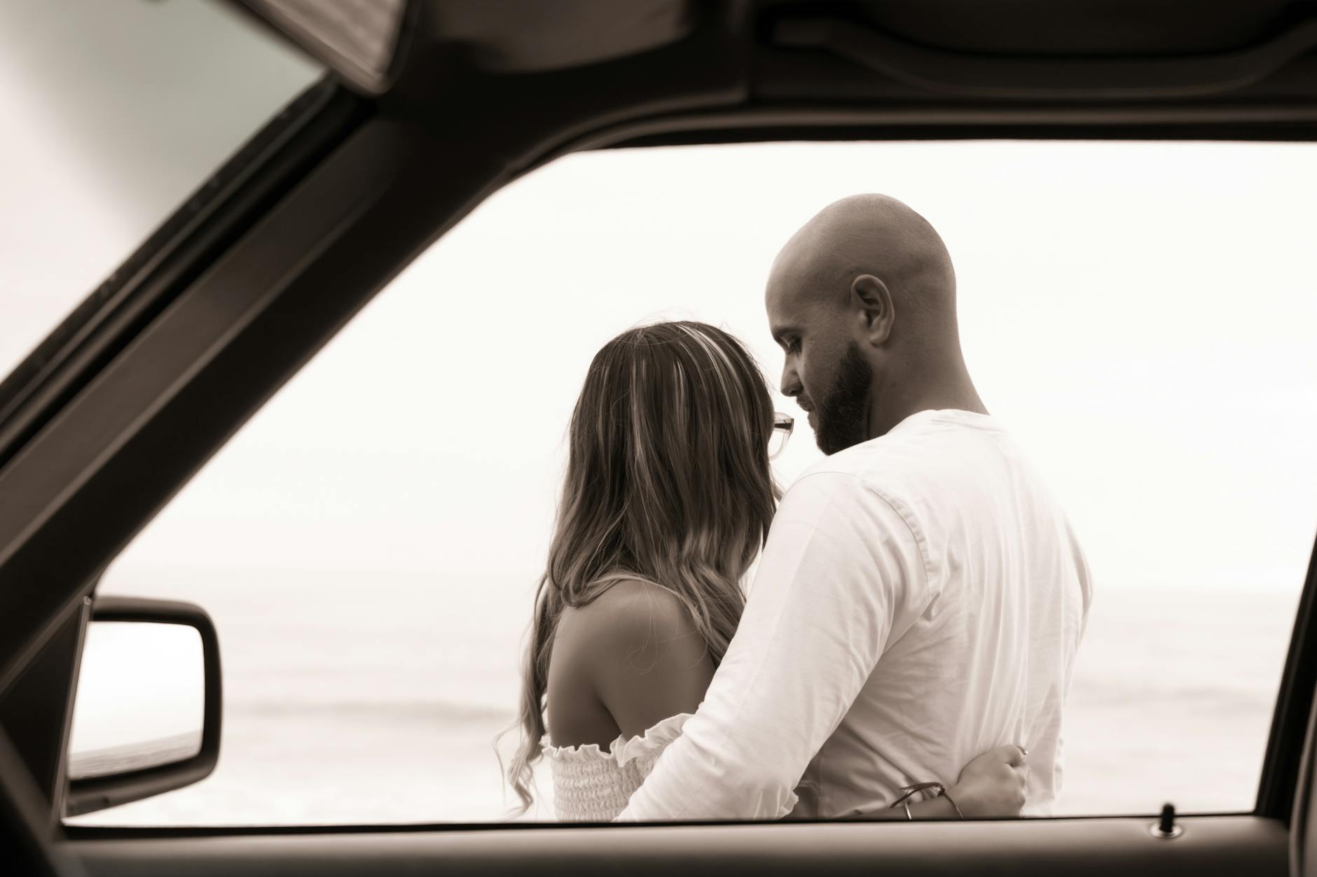 A romantic moment captured between a couple embracing near the ocean through a car window.