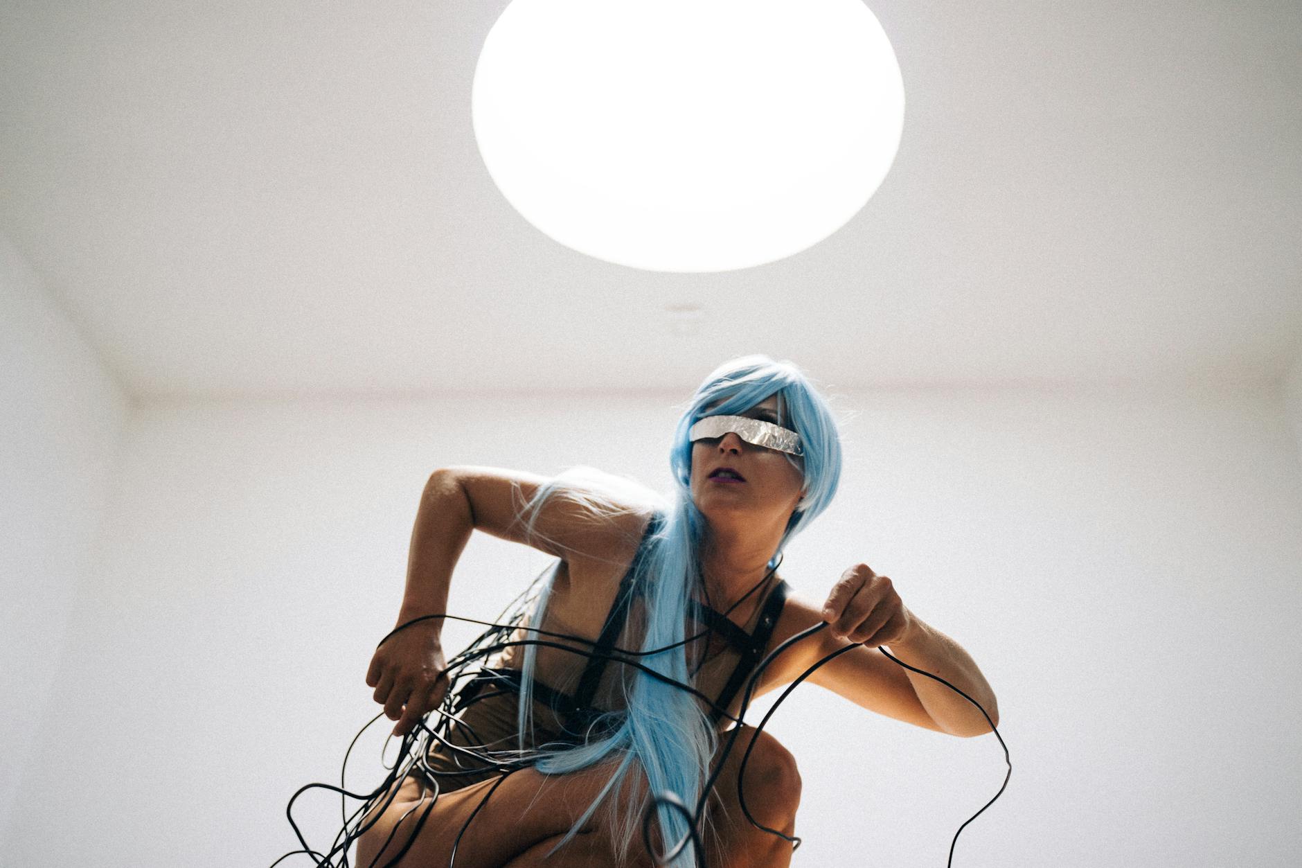 A striking pose of a woman in futuristic cyberpunk attire and blue hair in a minimalistic room.