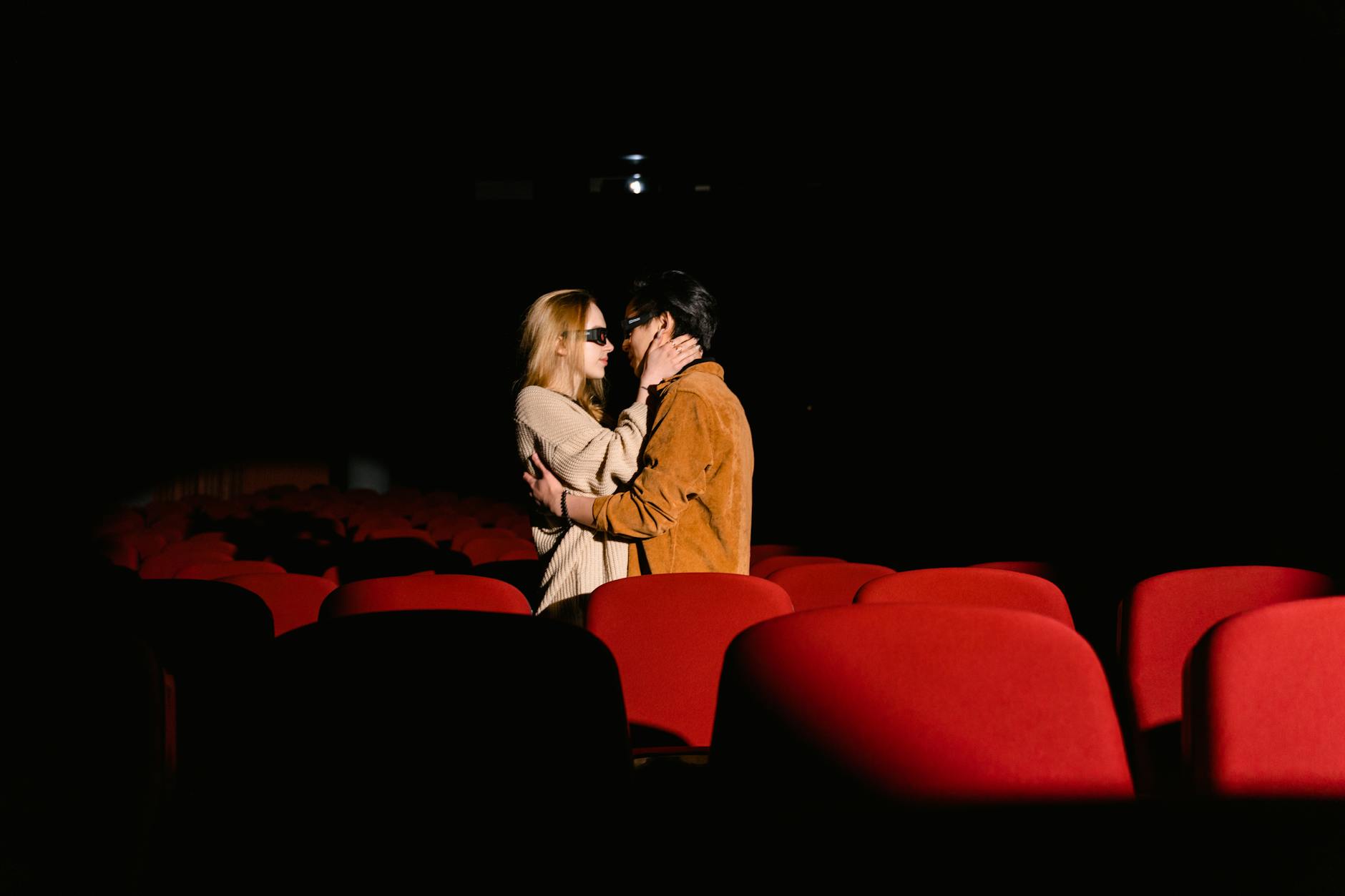 Couple sharing a tender moment in an empty movie theater with red seats.