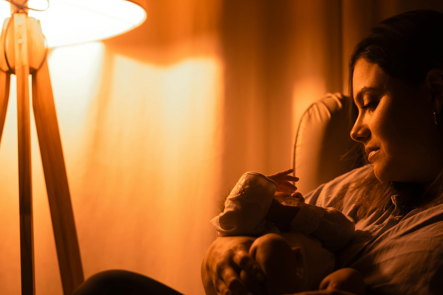 Mother softly gazes at her sleeping baby under warm lamplight, creating a serene and intimate atmosphere.