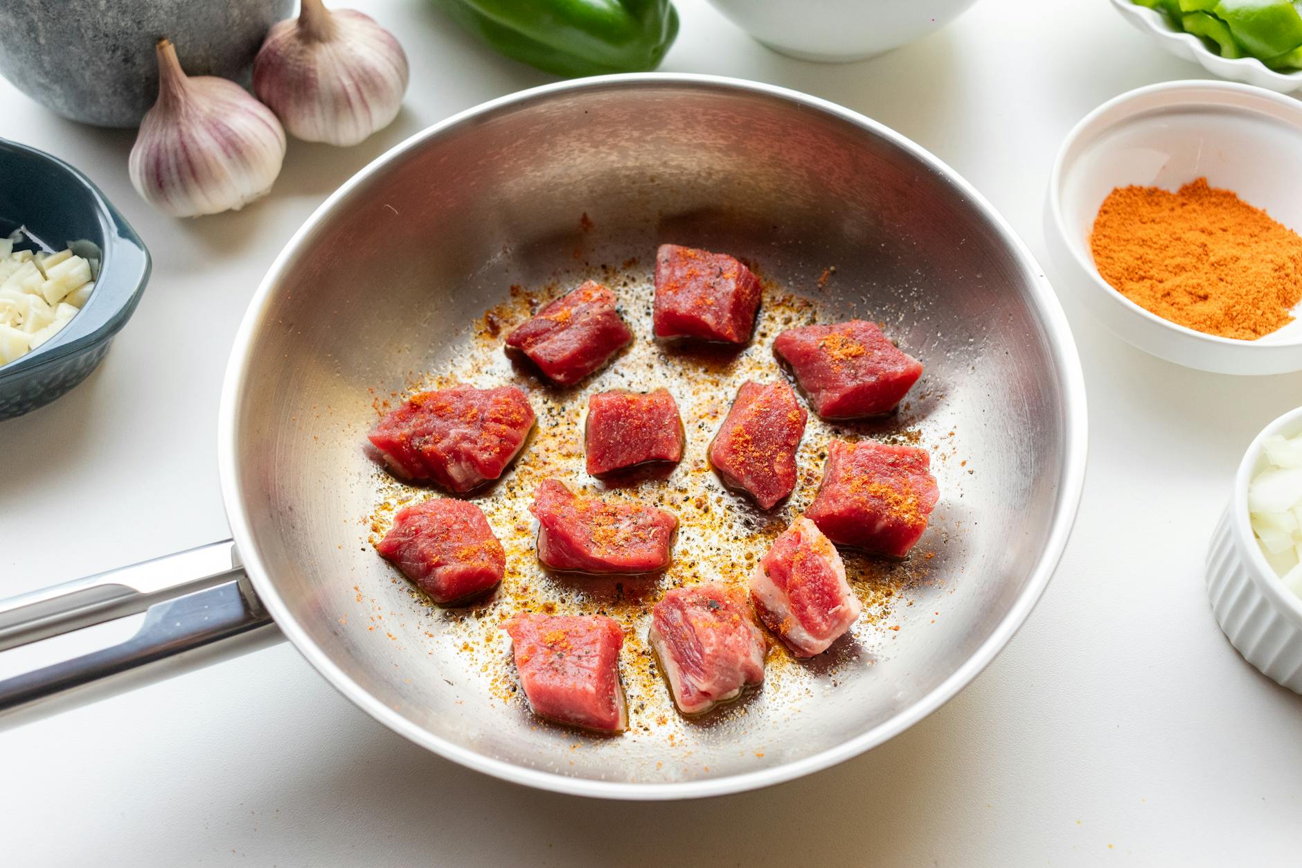Chunks of raw meat spiced and seared in a skillet surrounded by fresh ingredients.