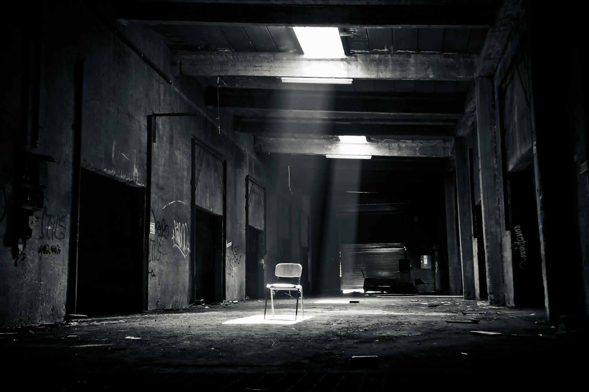 A haunting black and white photo of an abandoned warehouse with a solitary chair illuminated by sunlight through a window.