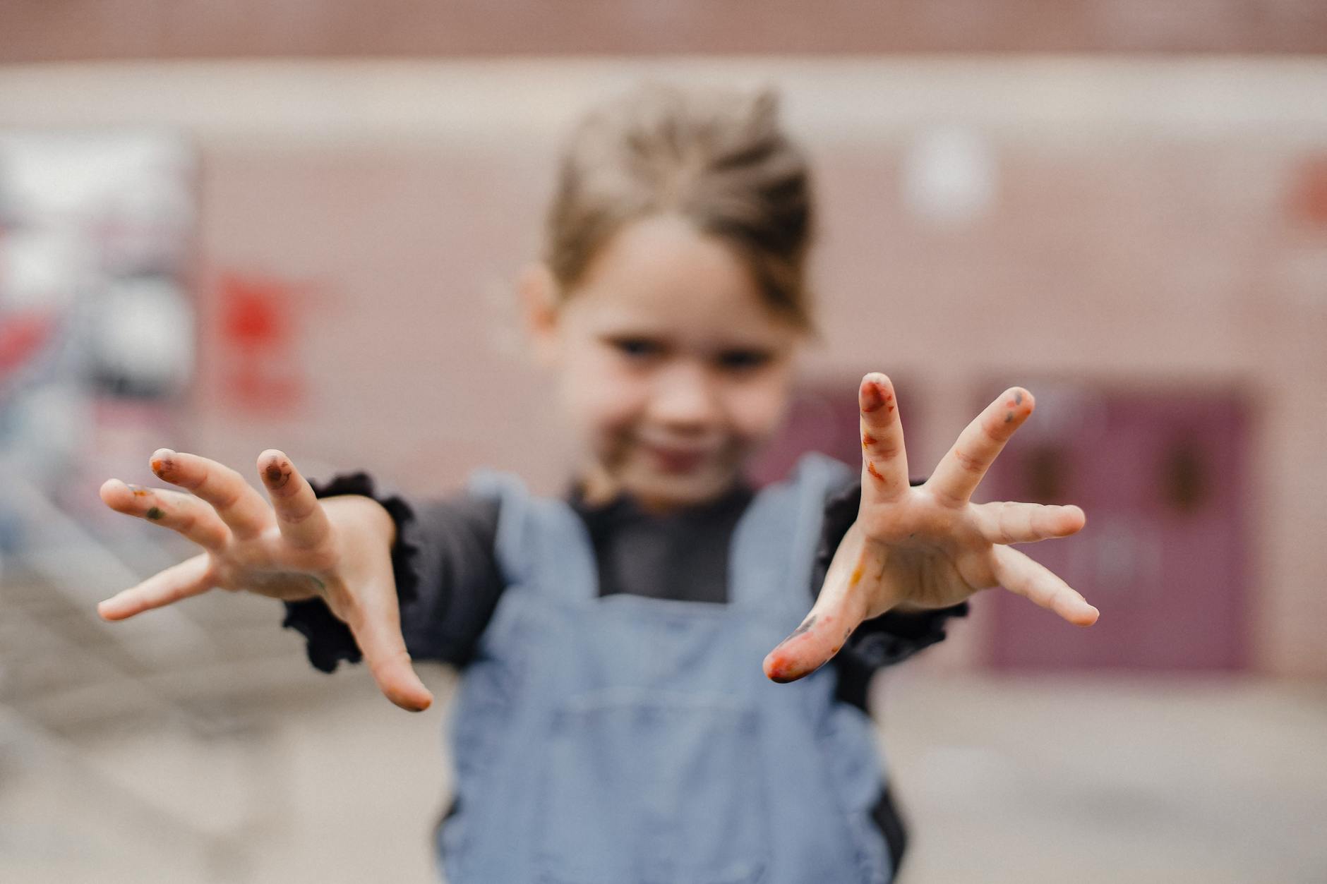 Adorable little girl in casual clothes with painted fingers outstretching arms towards camera while drawing in kindergarten