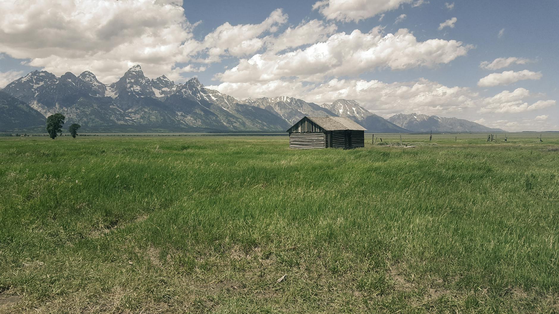 Rustic cabin in lush green field with Teton Mountain Range under a cloudy sky in Wyoming.