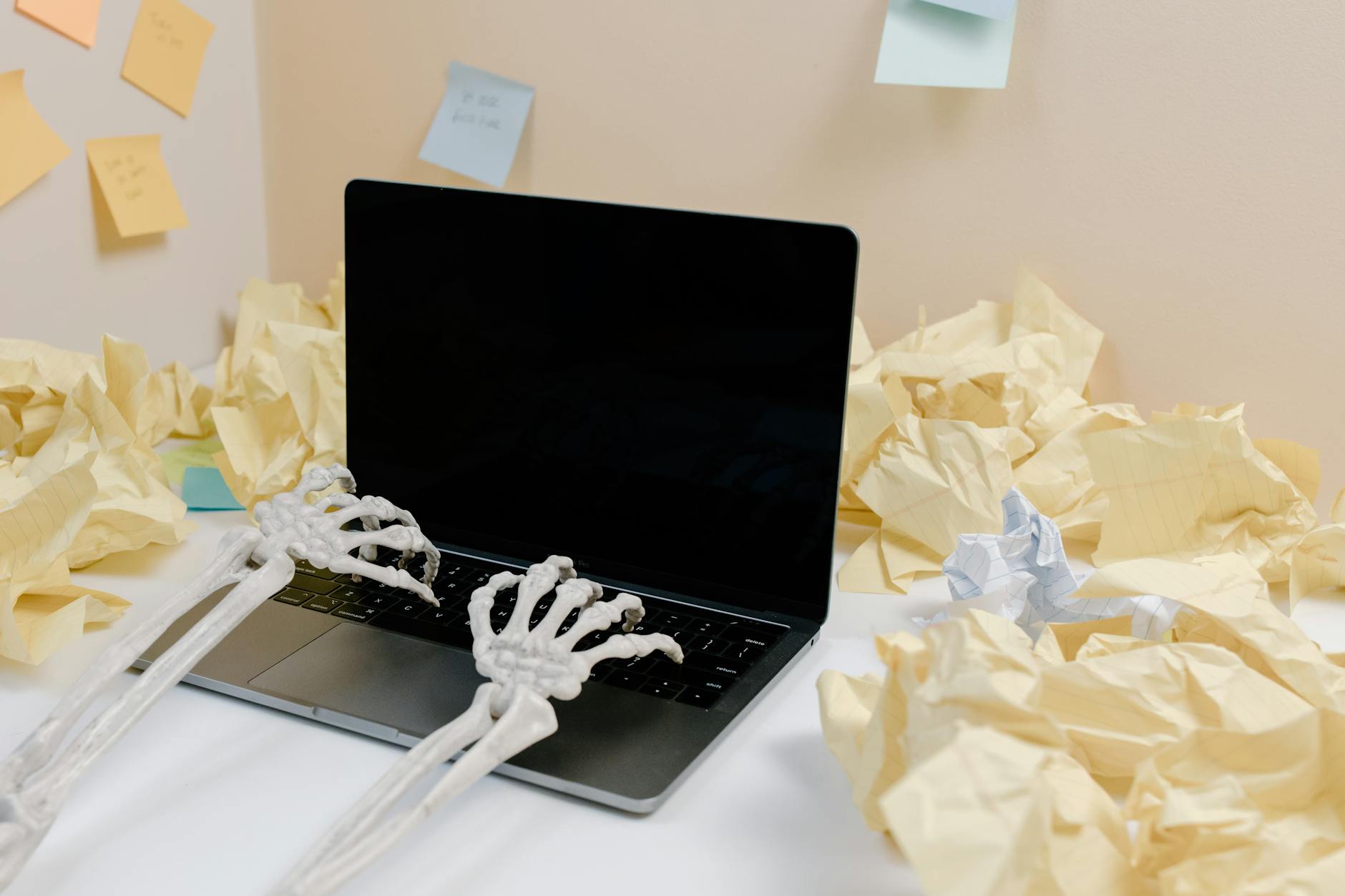 A humorous scene with skeleton hands typing on a laptop amid crumpled paper and sticky notes.