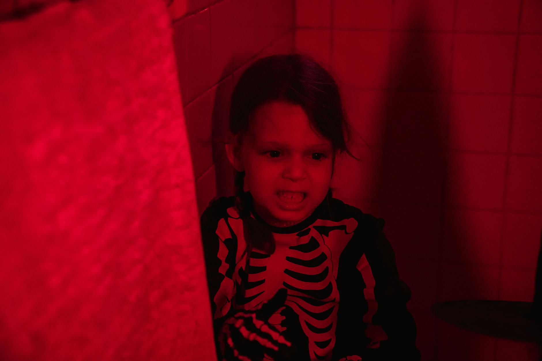 Child in a skeleton costume in a red-illuminated bathroom appears playful and spooky.