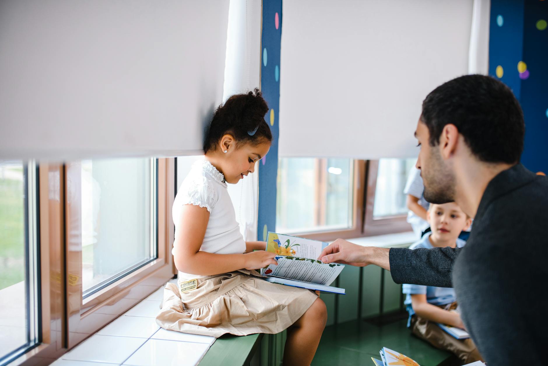 A teacher helps a young girl read a book with fellow students in a classroom setting.