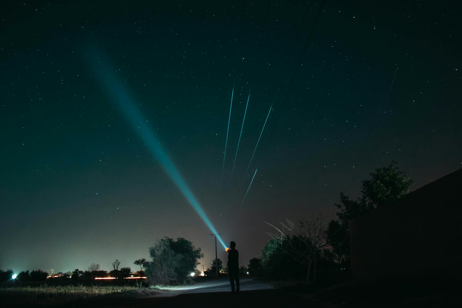 A man stands in a rural area, illuminating the night sky with a flashlight, under a starlit sky.