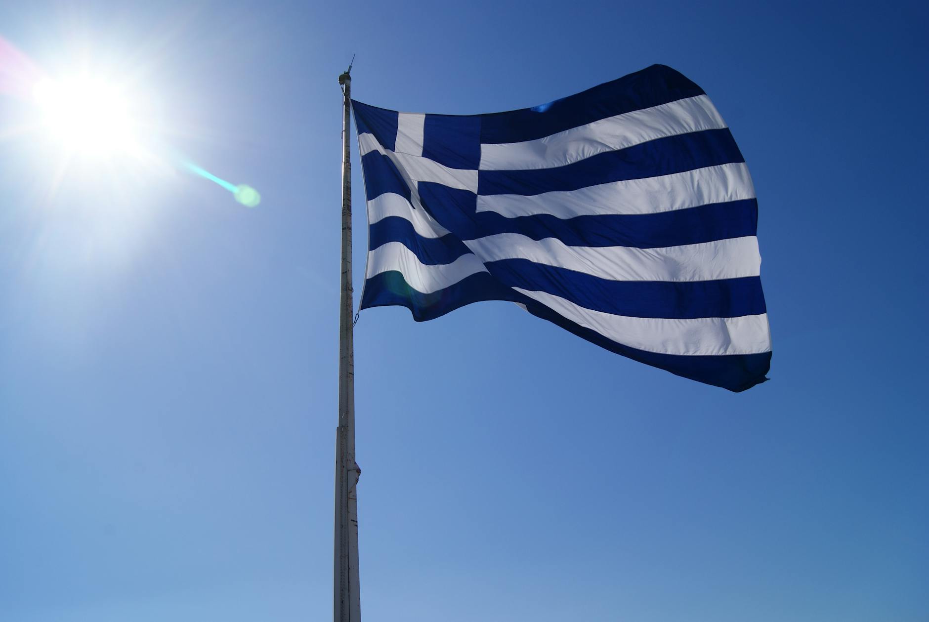 Greece's national flag waving against a clear blue sky and bright sun.
