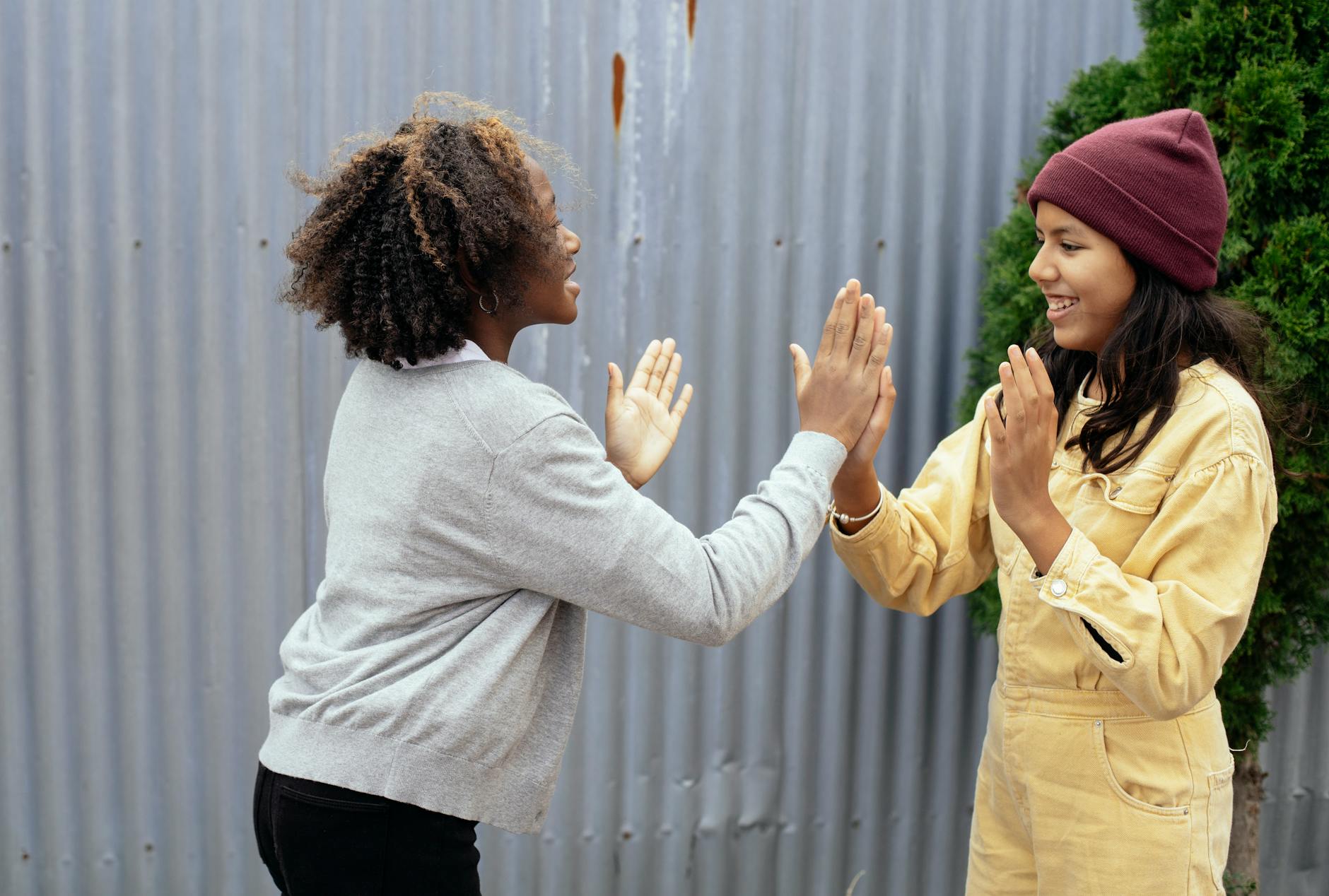 Side view of happy multiracial schoolgirls standing near metal fence while playing game with hands and looking at each other