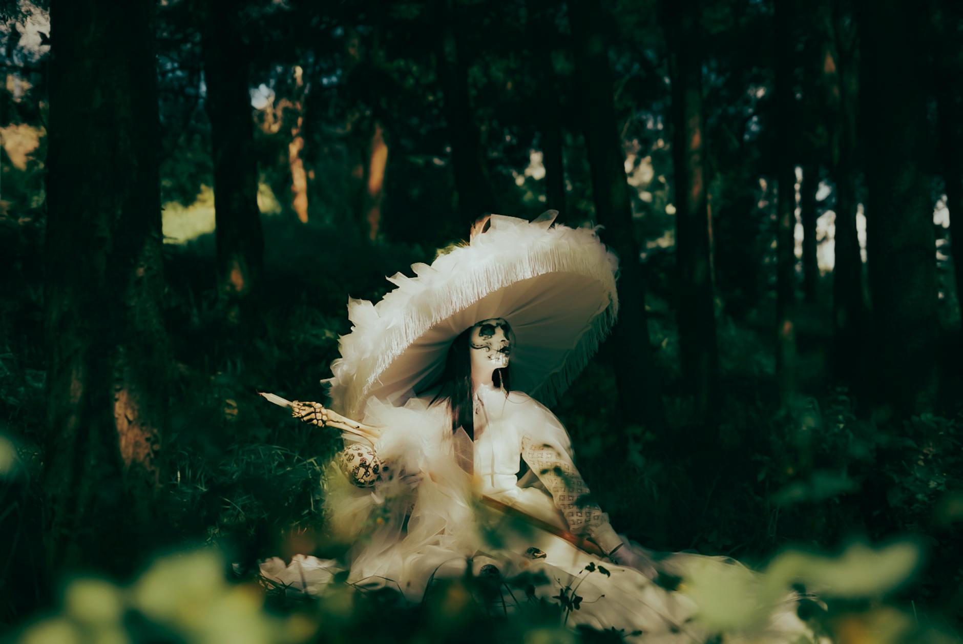 A woman with skeletal makeup wearing a large feathered hat in a mystical forest setting.