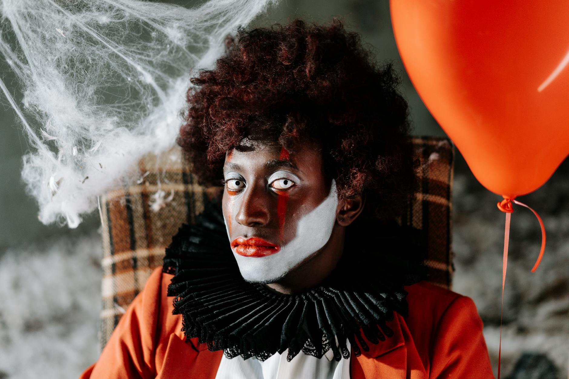 Spooky clown face with dramatic makeup, holding a red balloon amidst cobwebs. Halloween vibe.