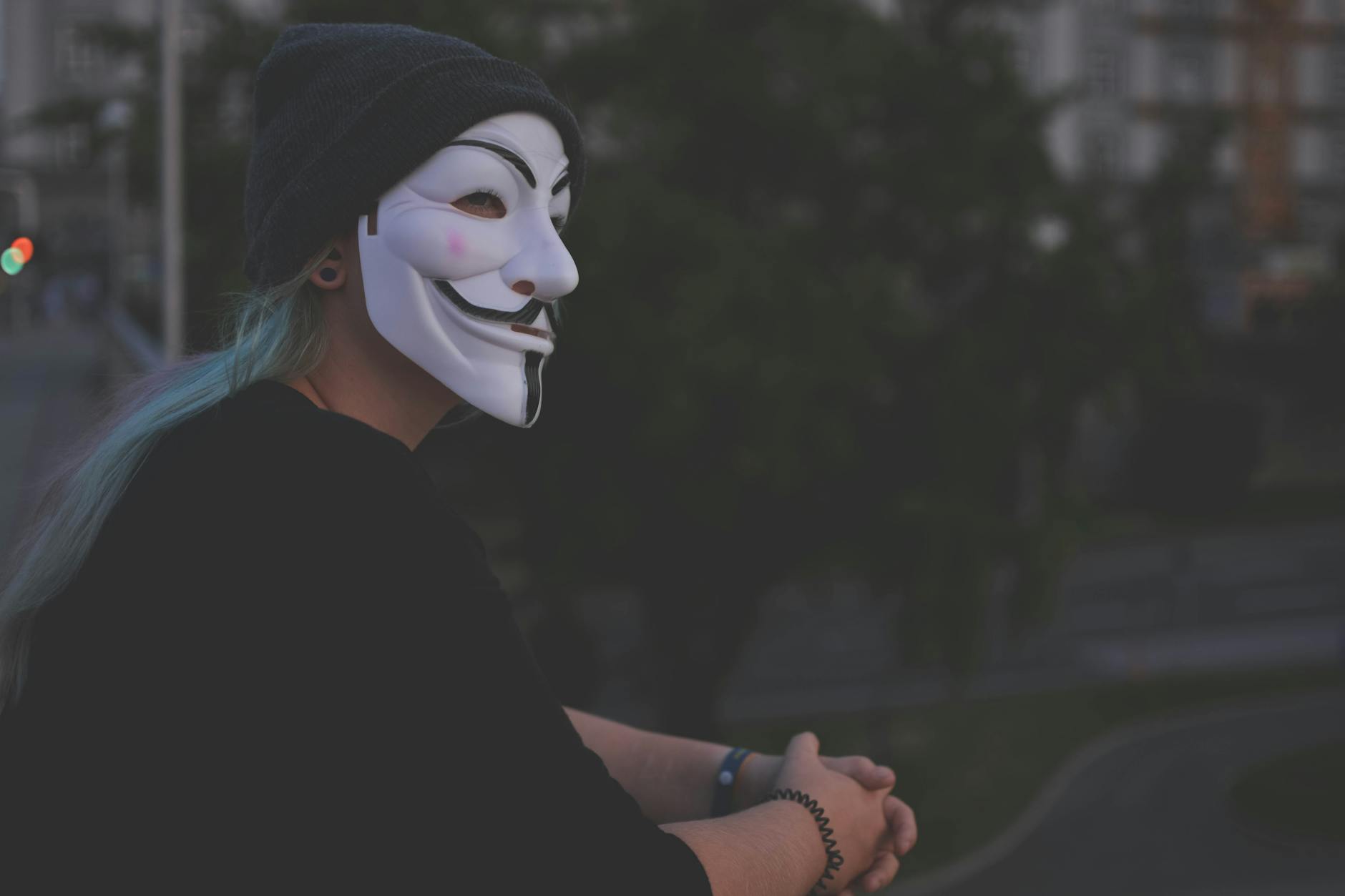A person wearing a Guy Fawkes mask stands on a balcony during twilight, creating an enigmatic scene.