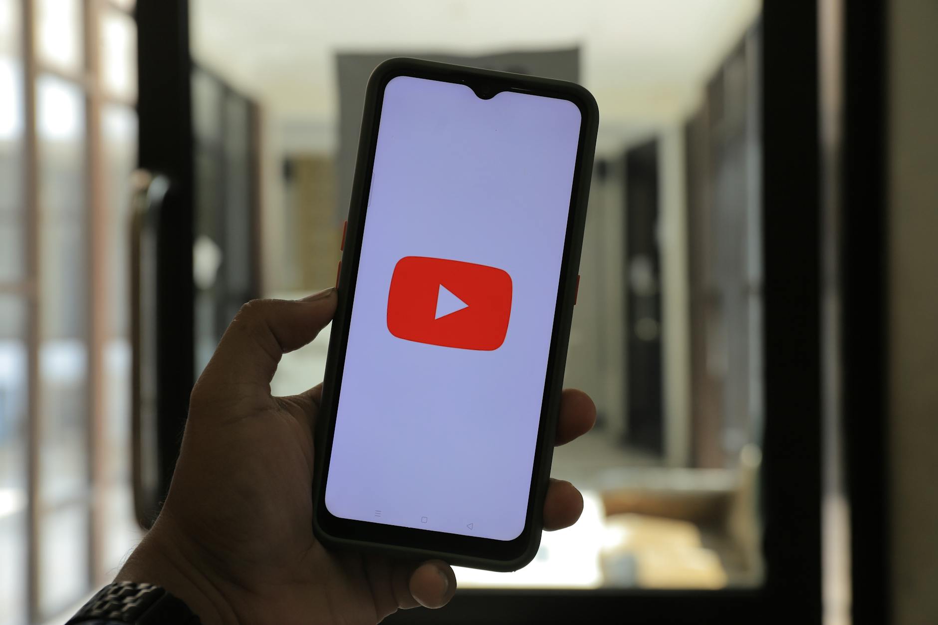 Close-up of a hand holding a smartphone displaying the YouTube logo in a well-lit room.