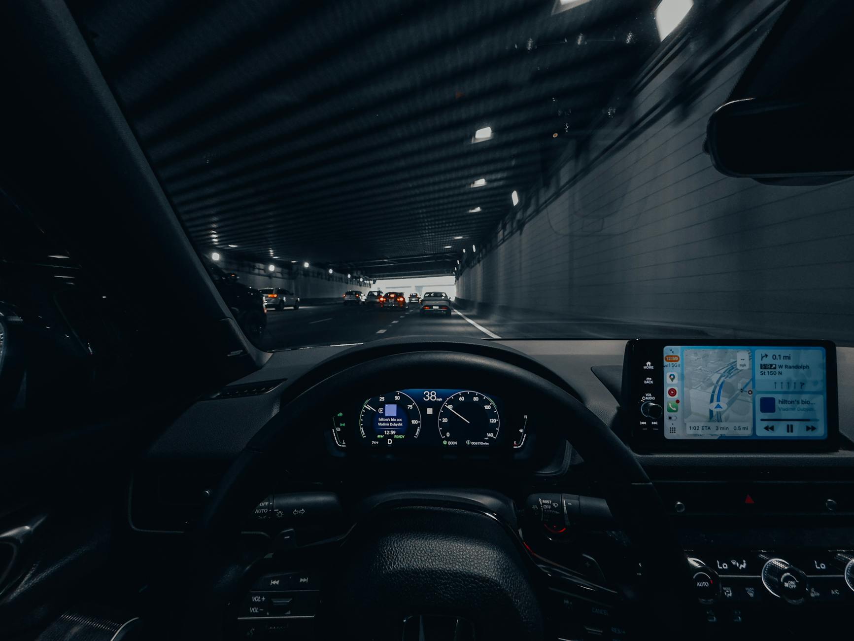 View from car dashboard driving through an illuminated city tunnel at night.