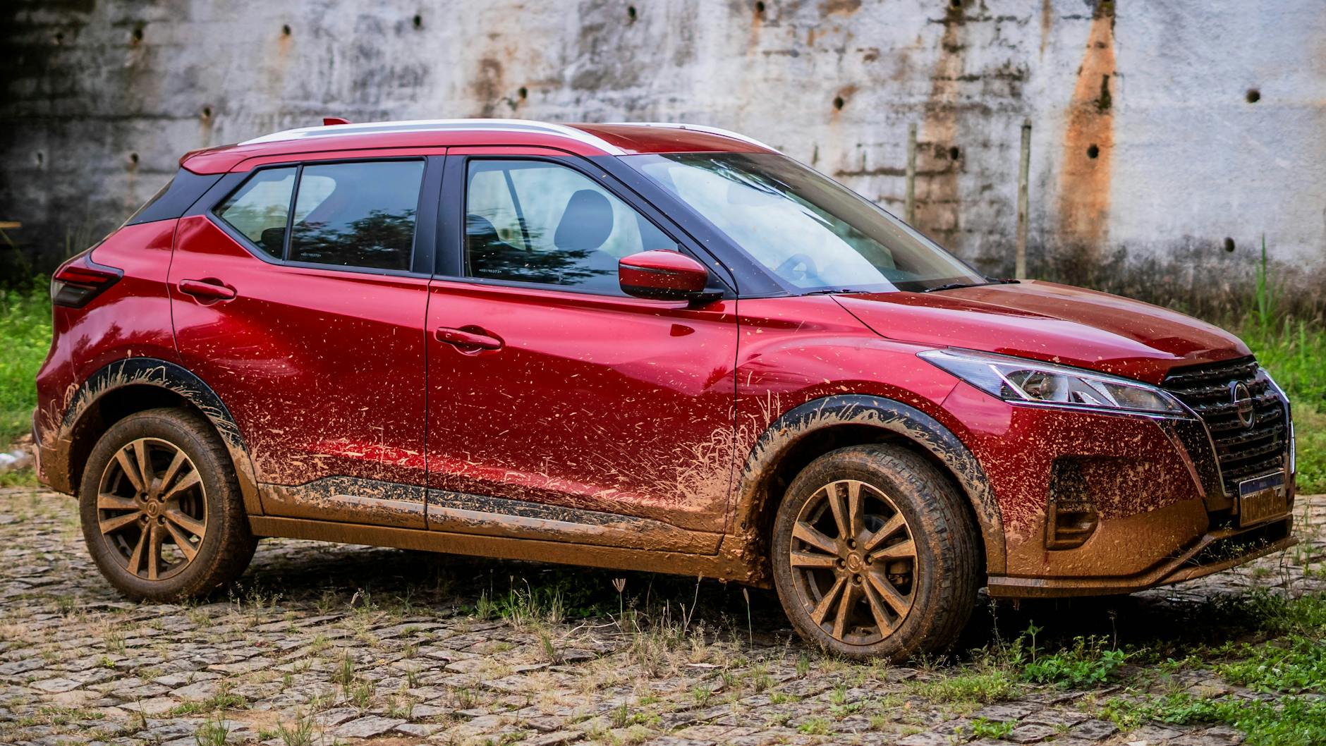 A red Nissan SUV with mud splatters seen parked on cobblestone.